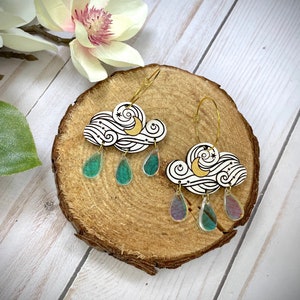 Puede incluir: Un par de pendientes de aro dorados con un dije de nube blanca y dijes de lágrima iridiscentes. El dije de nube presenta una luna creciente y estrellas.