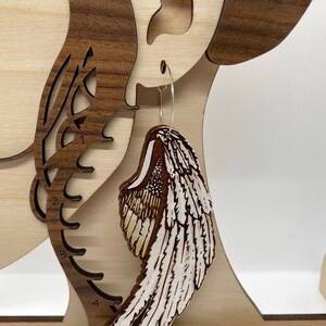 Pu&ograve; includere: Un paio di orecchini in legno a forma di ali d'angelo. Le ali sono dipinte di bianco con accenti marroni e hanno un design di piume dettagliato.