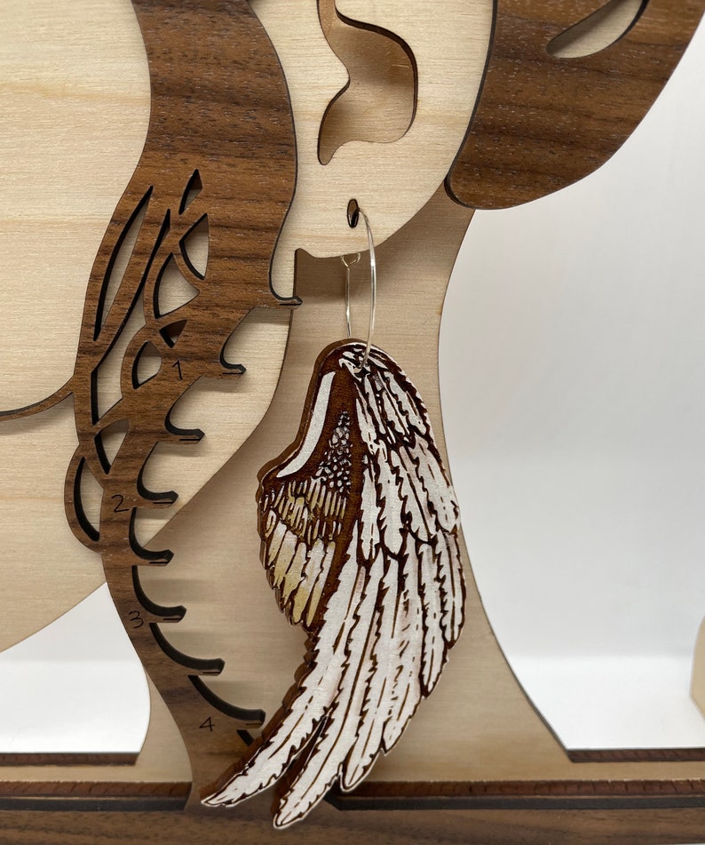 Pu&ograve; includere: Un paio di orecchini in legno a forma di ali d'angelo. Le ali sono dipinte di bianco con accenti marroni e hanno un design di piume dettagliato.