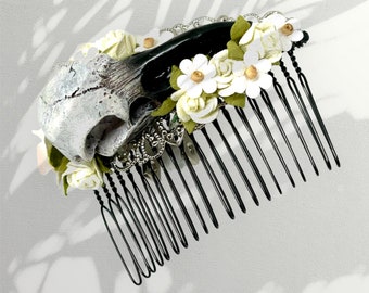May include: A black hair comb with a white skull and floral accents. The skull is made of resin and has a detailed texture. The flowers are white and green and are made of fabric.