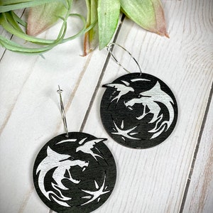 Puede incluir: Un par de pendientes de madera negra con una silueta blanca de dos lobos enfrentados. Los pendientes cuelgan de aros plateados.