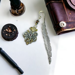 Könnte beinhalten: Ein silbernes Lesezeichen mit einem Federdesign und einem Steampunk-Zahnrad-Anhänger mit einem roten Juwel. Der Anhänger ist an einer silbernen Kette mit Perlen befestigt.