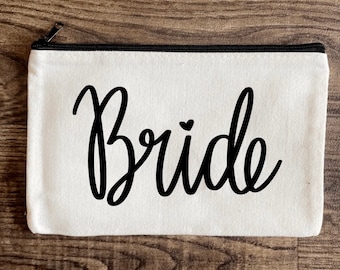 May include: White canvas zippered pouch with black cursive text that says "Bride" with a small heart above the "i".