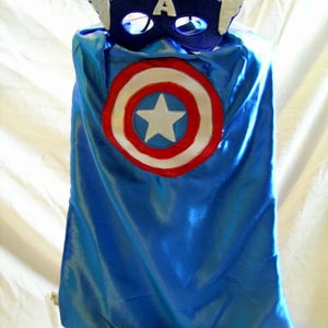May include: A blue satin cape with a red, white, and blue circle design in the center. The cape is topped with a blue felt mask with a white felt star and the letter "A" on the front.
