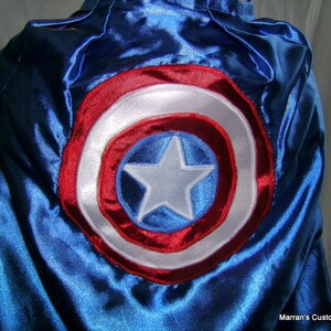 May include: A blue satin cape with a red, white, and blue embroidered circle design. The circle features a white star in the center.