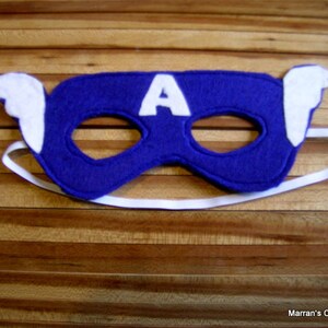 May include: A blue felt mask with a white felt "A" on the front and white felt wings on the sides. The mask has elastic straps.