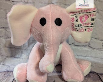 Op de afbeelding: Een roze pluche olifant met zwarte ogen en grote oren. De olifant zit op een houten oppervlak. Eén oor heeft geborduurde tekst en een voetafdrukontwerp. Een zacht speelgoed.