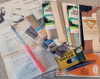 May include: A collection of vintage sewing supplies, including bias tape, seam binding, tailor's chalk, tracing paper, and a tracing wheel. The supplies are arranged on a wooden surface.