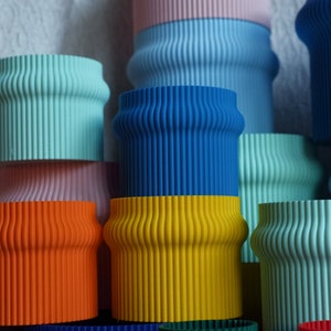 May include: A stack of colorful, ribbed, cylindrical planters in various shades of blue, green, yellow, orange, and pink. The planters are made of a smooth, textured material.