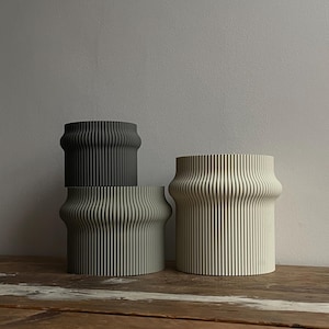 May include: Three ceramic planters with a ribbed design. The planters are in shades of grey and beige. The planters are sitting on a wooden surface.