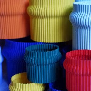 May include: A stack of colorful, ribbed, cylindrical planters in various shades of blue, green, yellow, orange, and red. The planters are made of a smooth, plastic-like material.