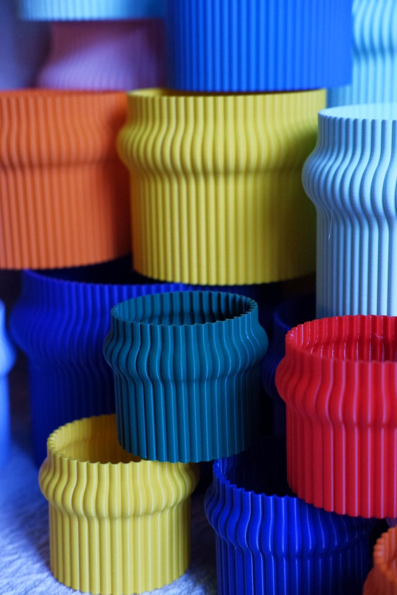 May include: A stack of colorful, ribbed, cylindrical planters in various shades of blue, green, yellow, orange, and red. The planters are made of a smooth, plastic-like material.