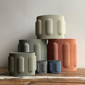 May include: Set of five cylindrical planters in various sizes. The planters are made of a textured material and have a ribbed design. The colours are green, orange, and blue.