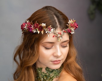 May include: A woman with long, wavy red hair wears a floral crown with red and gold accents. She is wearing a green and gold sequined dress with a plunging neckline.
