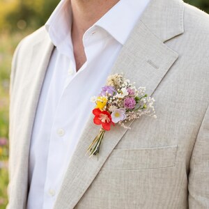 Op de afbeelding: Lichtbeige blazer over een wit overhemd, versierd met een kleurrijke bloemenknop. De knop bevat rode, gele en paarse bloemen. De blazer is gemaakt van een textuurstof, tegen een achtergrond van wilde bloemen.