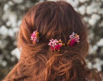 May include: A woman with long, curly, auburn hair has three small flower hair clips in her hair. The clips are pink, yellow, and red.