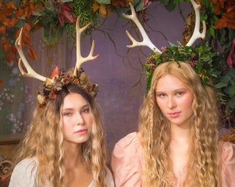 May include: Two women wearing floral crowns with antlers, one in a white dress and the other in a pink dress, pose in front of a backdrop of greenery.