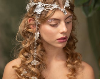 May include: A woman with long, curly blonde hair wears a white lace off-the-shoulder top and a delicate silver floral crown with dangling crystals.