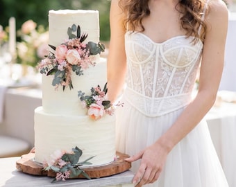 May include: A three-tiered white wedding cake decorated with pink and peach flowers and greenery. The cake sits on a wooden slab. The person in the image is wearing a white dress and a floral crown. The background is a garden setting.