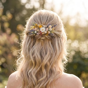 Op de afbeelding: Een haarkam versierd met een arrangement van gedroogde bloemen in tinten geel, wit, roze en paars. Het bloemstuk is bevestigd in blond haar dat half opgestoken is met losse golven die langs de rug naar beneden vallen.