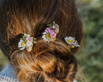 May include: A woman with long brown hair has a bun with small white and pink flowers in her hair.