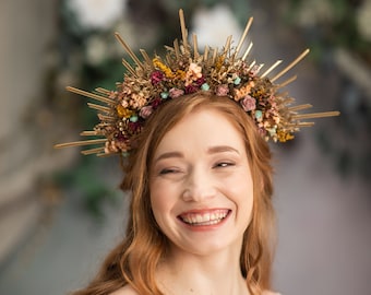 May include: A woman with long, curly red hair wears a gold floral crown with dried flowers. She is smiling and looking to the side. She is wearing a white, off-the-shoulder dress.