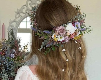 Könnte beinhalten: Eine Person mit langen, welligen blonden Haaren, die ein weißes Kleid und einen Blumenkranz trägt. Sie hält einen Strauß bunter Blumen. Der Hintergrund umfasst einen Spiegel und Kerzen, was auf eine Hochzeit oder einen besonderen Anlass hindeutet.