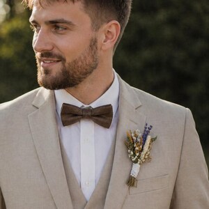 May include: A beige linen suit with a matching vest and a white dress shirt. The suit features a brown bow tie and a small floral boutonniere. The ensemble is suitable for a wedding or formal event.