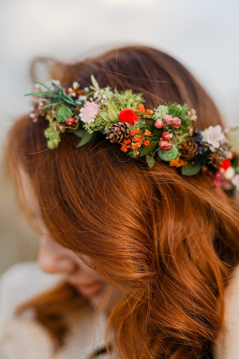 May include: A woman with long, red hair wearing a floral crown with red berries, pine cones, and green leaves.