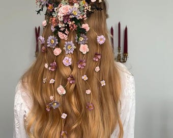 May include: A woman with long, wavy, light brown hair wearing a white dress with a floral hairpiece. The hairpiece is made of pink, purple, and white flowers and is cascading down the back of her hair.