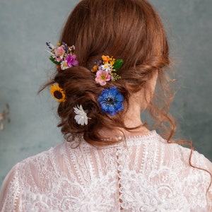 May include: A woman with long, curly red hair wearing a white lace dress with a low back. Her hair is styled in a bun with a variety of colorful flowers, including blue, yellow, pink, and purple.