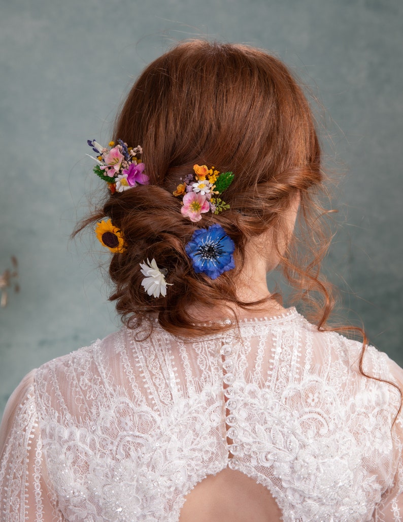 May include: A woman with long, curly red hair wearing a white lace dress with a low back. Her hair is styled in a bun with a variety of colorful flowers, including blue, yellow, pink, and purple.