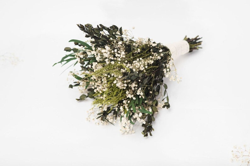 May include: A bouquet of dried flowers with dark green leaves, tiny white flowers, and light green sprigs. The stems are bound with a cream-coloured ribbon, creating a natural and elegant arrangement.
