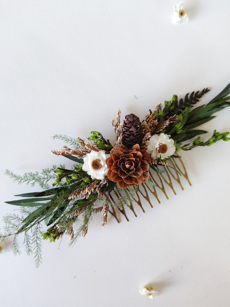 May include: A gold hair comb decorated with dried flowers and pine cones. The flowers are white and the pine cones are brown.