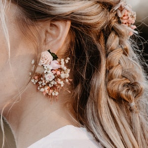 Könnte beinhalten: Ein Paar Blumenohrringe mit rosa und weißen Blüten und Grün. Die Ohrringe werden von einer Person mit langen blonden Haaren getragen.
