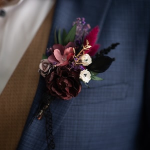 May include: A dark blue suit jacket with a buttonhole featuring burgundy, pink, and white flowers, purple and green foliage, and black feathers.