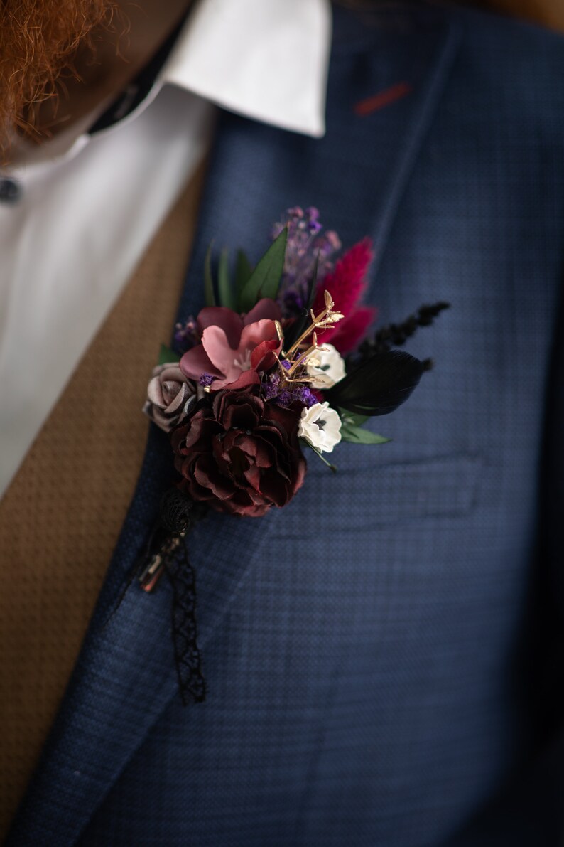 May include: A dark blue suit jacket with a buttonhole featuring burgundy, pink, and white flowers, purple and green foliage, and black feathers.