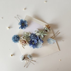 K&ouml;nnte beinhalten: Eine Auswahl an floralen Haarnadeln und kleinen Blumen, die auf einer wei&szlig;en Oberfl&auml;che angeordnet sind. Die Haarnadeln weisen blaue, wei&szlig;e und rosa Stoffblumen auf. Die Anordnung umfasst einen offenen Umschlag mit einer leeren wei&szlig;en Karte.