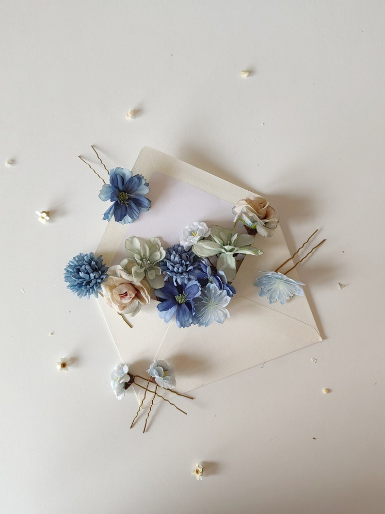 K&ouml;nnte beinhalten: Eine Auswahl an floralen Haarnadeln und kleinen Blumen, die auf einer wei&szlig;en Oberfl&auml;che angeordnet sind. Die Haarnadeln weisen blaue, wei&szlig;e und rosa Stoffblumen auf. Die Anordnung umfasst einen offenen Umschlag mit einer leeren wei&szlig;en Karte.