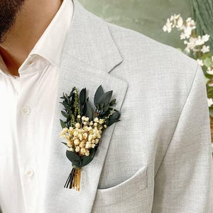 May include: A light grey blazer with a white dress shirt and a floral buttonhole. The buttonhole features small white flowers, dark green leaves, and gold-coloured stems, tied with green twine. The blazer has a pocket on the left side.