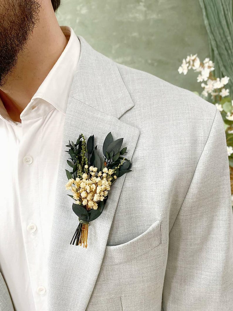 May include: A light grey blazer with a white dress shirt and a floral buttonhole. The buttonhole features small white flowers, dark green leaves, and gold-coloured stems, tied with green twine. The blazer has a pocket on the left side.