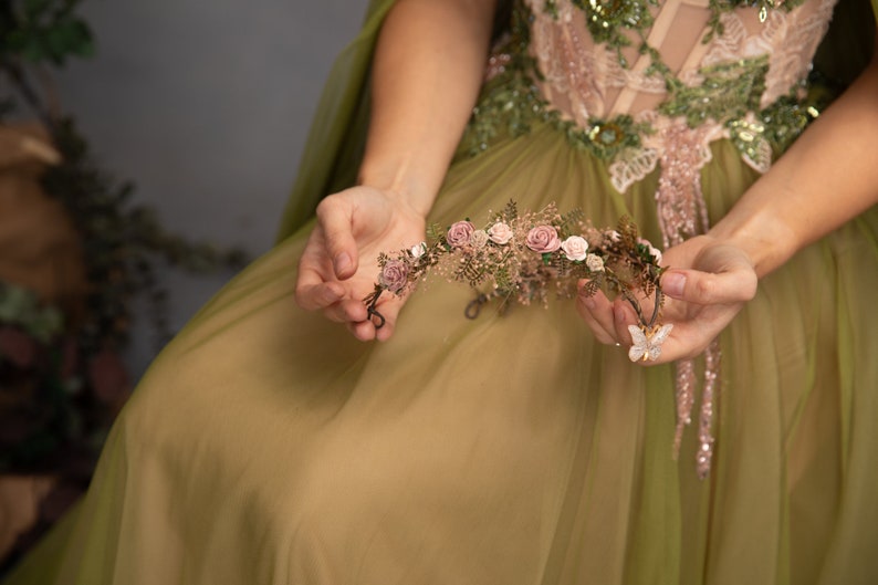 Peut inclure: Une couronne florale d&eacute;licate avec des fleurs roses et des feuilles vertes, tenue dans les mains d'une personne portant une robe verte.