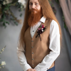 May include: A man with long red hair and a beard is wearing a brown waistcoat with a white shirt and blue trousers. He has a small bouquet of flowers pinned to his waistcoat.