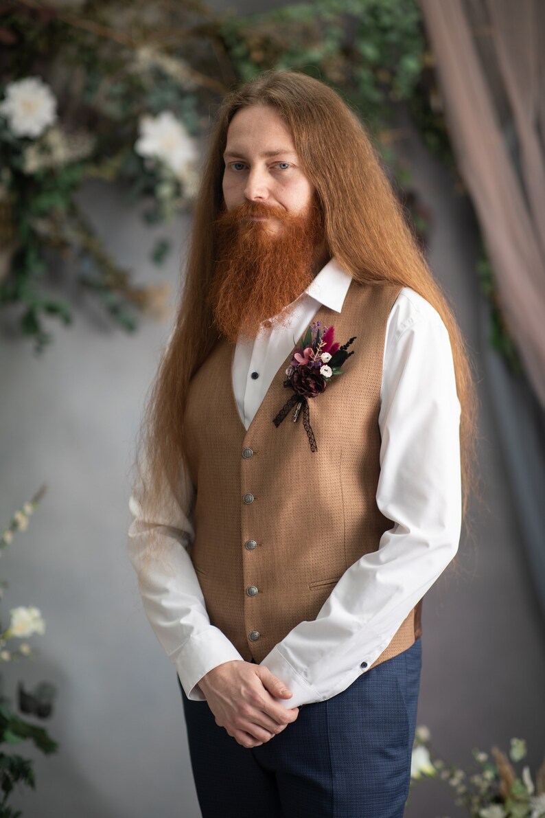 May include: A man with long red hair and a beard is wearing a brown waistcoat with a white shirt and blue trousers. He has a small bouquet of flowers pinned to his waistcoat.