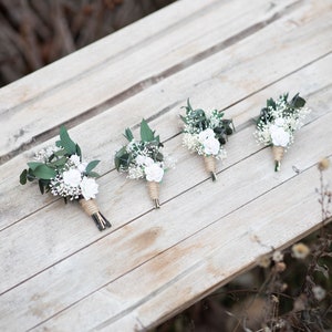 Puede incluir: Cuatro boutonnieres con rosas blancas, paniculata y vegetaci&oacute;n sobre una superficie de madera envejecida. Los tallos est&aacute;n envueltos con cuerda. Son arreglos florales para bodas u ocasiones especiales.