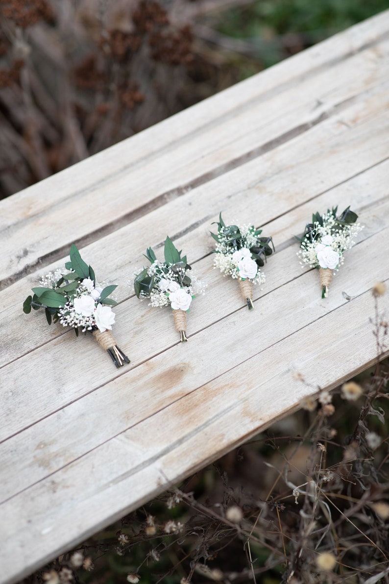 Puede incluir: Cuatro boutonnieres con rosas blancas, paniculata y vegetaci&oacute;n sobre una superficie de madera envejecida. Los tallos est&aacute;n envueltos con cuerda. Son arreglos florales para bodas u ocasiones especiales.
