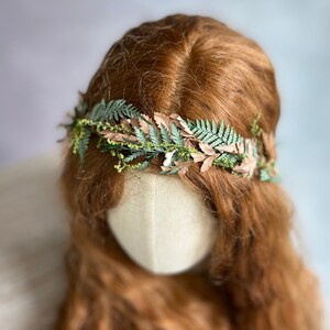 May include: A floral crown featuring green fern leaves and tan-colored leaves, accented with small yellow flowers. The crown is set against a backdrop of long, wavy auburn hair and a neutral-toned mannequin head, creating a natural, earthy aesthetic.