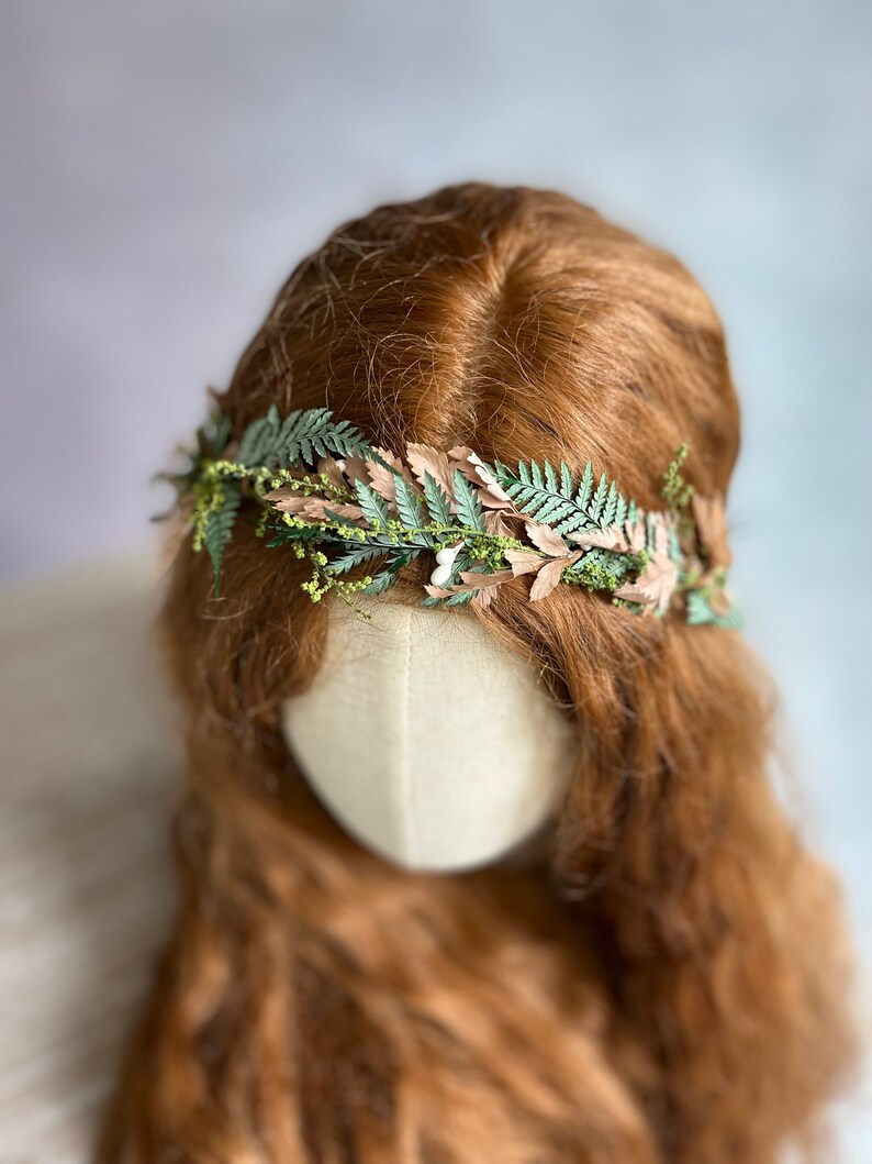 May include: A floral crown featuring green fern leaves and tan-colored leaves, accented with small yellow flowers. The crown is set against a backdrop of long, wavy auburn hair and a neutral-toned mannequin head, creating a natural, earthy aesthetic.