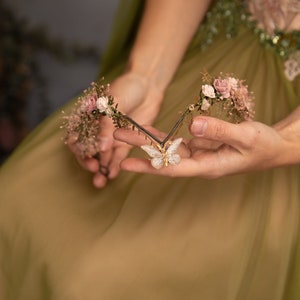 Peut inclure: Une couronne florale d&eacute;licate avec des fleurs roses et de la verdure, orn&eacute;e d'un charme papillon scintillant au centre.