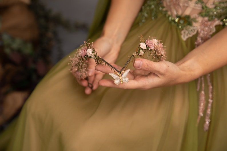 Peut inclure: Une couronne florale d&eacute;licate avec des fleurs roses et de la verdure, orn&eacute;e d'un charme papillon scintillant au centre.